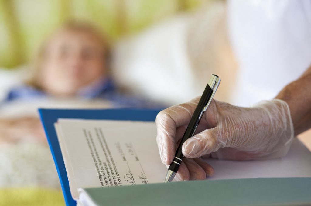 Care professional writing notes by bedside wearing surgical gloves