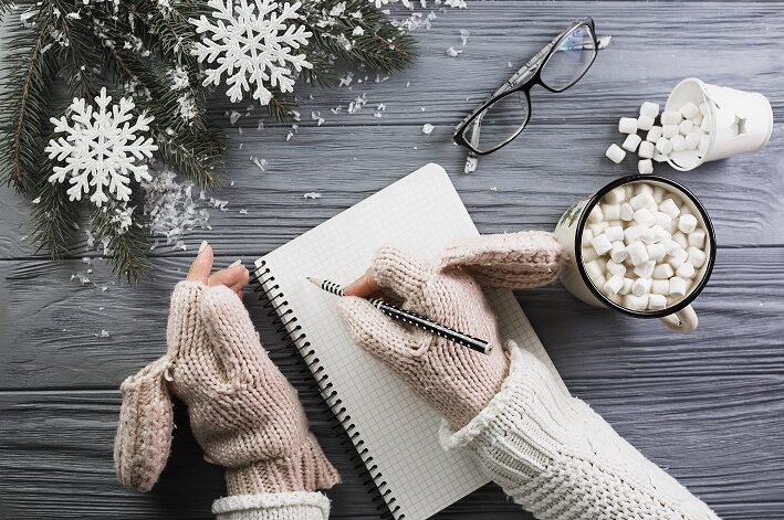 woman-mittens-writing-notebook-near-cup-with-marshmallow-eyeglasses